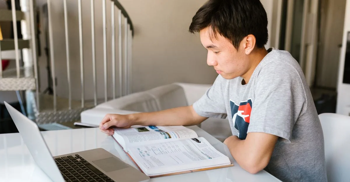 Un garçon adolescent se concentre sur ses devoirs en utilisant un ordinateur portable et un manuel à l'intérieur.