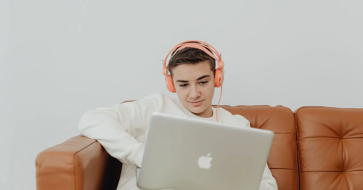 Jeune homme assis sur un canapé utilisant un ordinateur portable avec des écouteurs, profitant de la musique à l'intérieur.