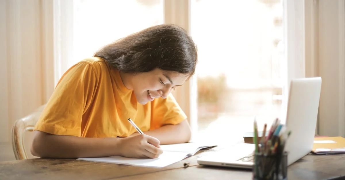 Jeune femme étudiant avec un ordinateur portable et un bloc-notes à la maison, souriante et concentrée.