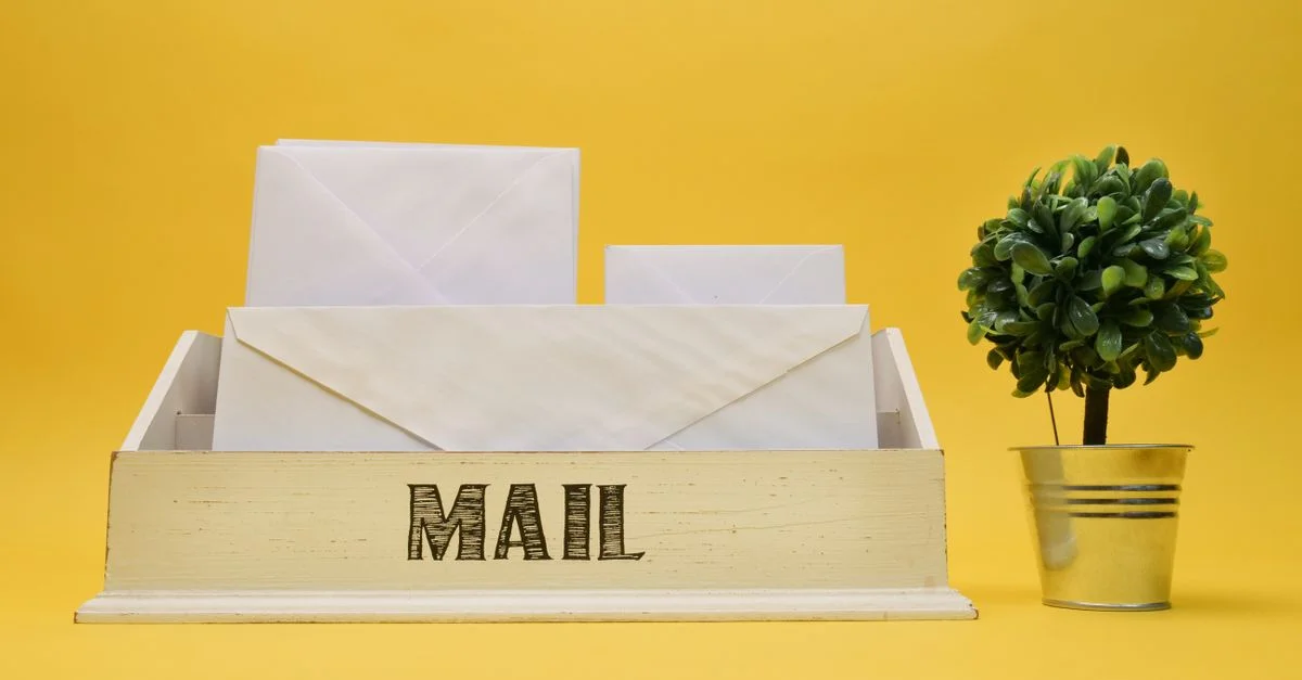 Un organisateur de courrier en bois avec des enveloppes blanches et une petite plante en pot sur un fond jaune.