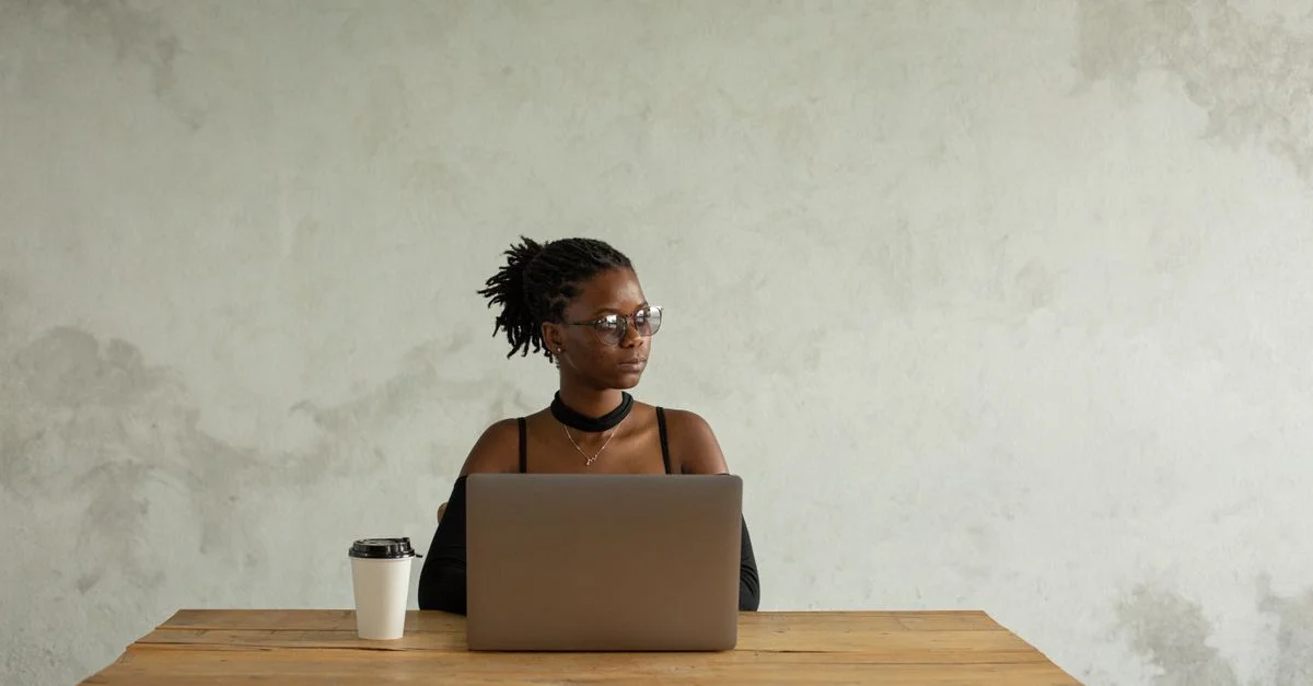 Une femme en robe noire avec des lunettes travaille sur un ordinateur portable à une table en bois.