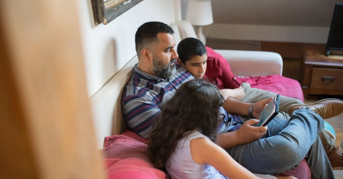 Un père et ses enfants se détendent sur un canapé en utilisant une tablette numérique dans un cadre domestique chaleureux.