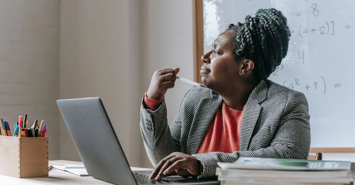 Une femme afro-américaine dans une pose réfléchie travaillant à un bureau avec un ordinateur portable et des fournitures de bureau.