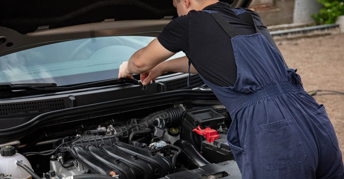 Un mécanicien en bleu de travail travaille sur un moteur de voiture en extérieur, mettant en avant ses compétences en entretien de véhicule.