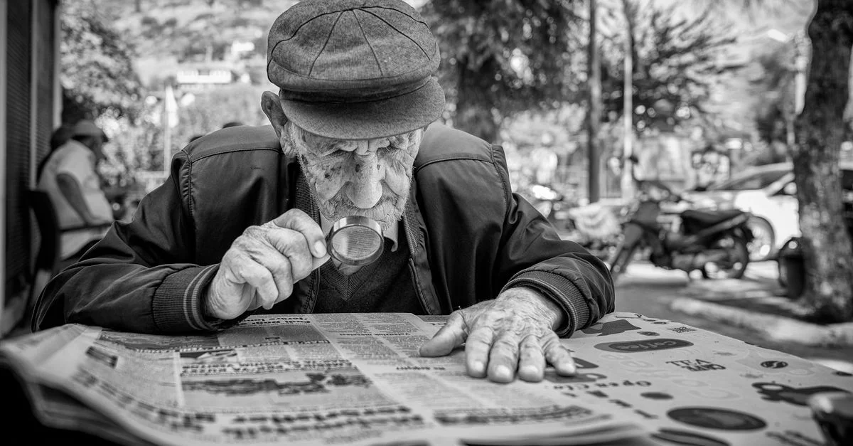 Un homme senior utilise une loupe pour lire un journal en plein air, mettant en avant le vieillissement et la concentration.